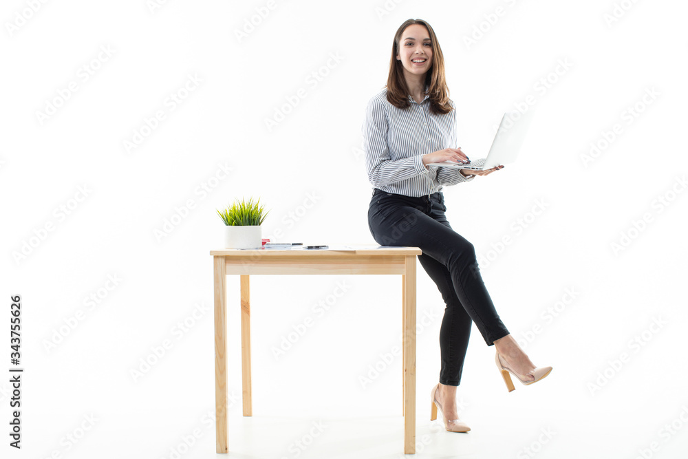 A girl in a good mood is sitting on a table and typing on a computer on a white background