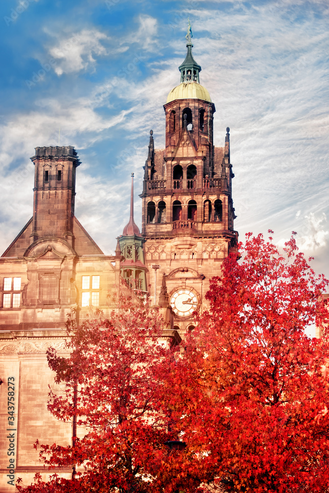 Obraz premium Yellow town hall with clock and green dome and red tree in front of it at the sunset against cloudy blue sky in the Autumn