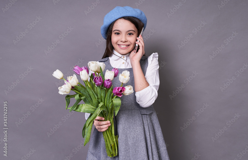 Beautiful cute Caucasian lady in a stylish blue hat in a beret with a phone holds tulips flowers in her hands on a gray background. Beautiful cute Caucasian lady in a stylish blue hat in a beret with a phone holds tulips flowers in her hands on a gray background.