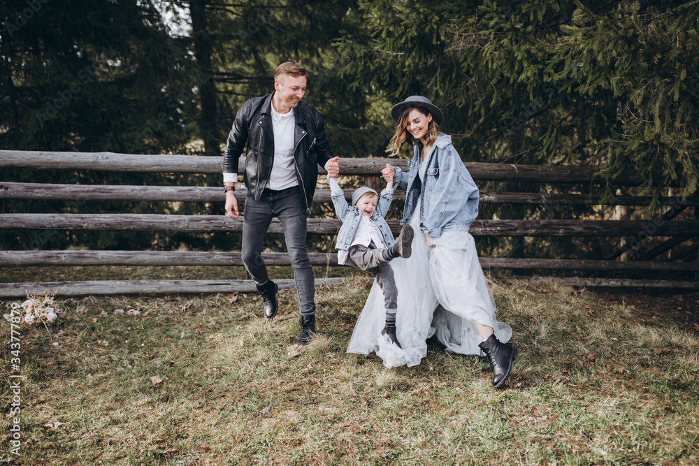 Fototapeta premium Stylish family in the mountains. A guy in a leather jacket, a girl in a wedding dress and hat run and play with his son against a background of green firs and a wooden fence