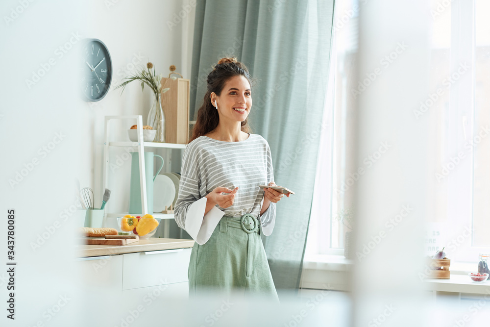Stylish young woman wearing casual outfit standing in kitchen taking part in online meeting using her smartphone and earphones Stylish young woman wearing casual outfit standing in kitchen taking part in online meeting using her smartphone and earphones
