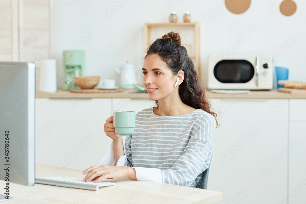 Horizontal medium portrait of young woman using her desktop computer and drinking morning coffee at home Horizontal medium portrait of young woman using her desktop computer and drinking morning coffee at home