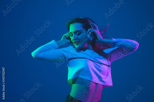 Listening to music. Caucasian young woman's portrait isolated on blue studio background in neon light. Beautiful female model. Concept of human emotions, facial expression, sales, ad, youth culture.