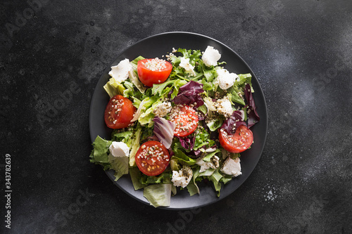 Healthy vegan salad in black dish with fresh tomato, greens, seeds hemp. Top view.