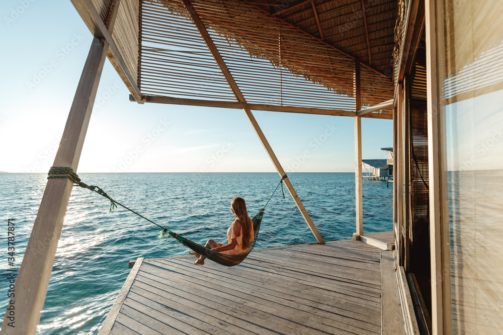 Happy smiling woman lies in hammock swing over the ocean. Back view woman  sitting in hammock on the beach in water bungalow.  Travel and vacation concept