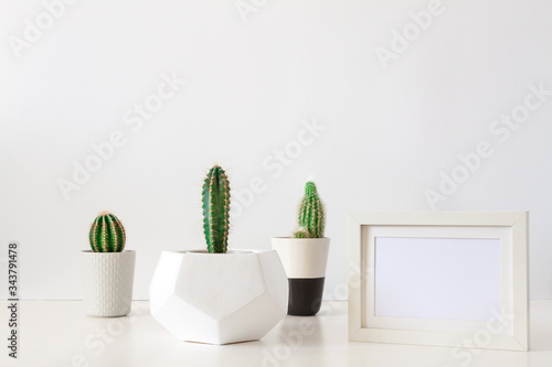 Desk on a white empty wall background with three cactuses in a geometric pot and a frame mockup. Copy space.