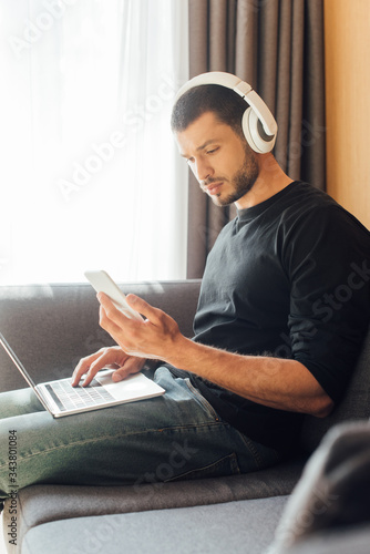 selective focus of freelancer in wireless headphones looking at smartphone near laptop