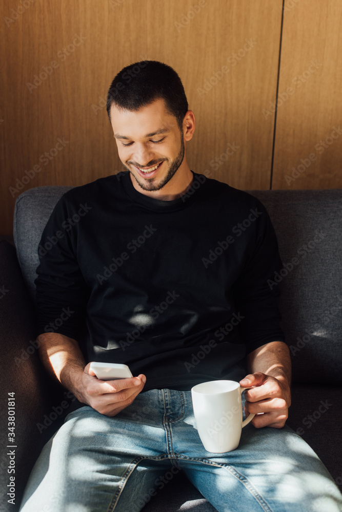 sunlight on bearded man smiling while using smartphone and holding cup in living room sunlight on bearded man smiling while using smartphone and holding cup in living room