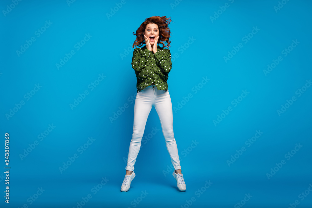 Full body photo of pretty crazy lady jumping high arms on cheekbones good news open mouth wear green dotted shirt white pants shoes isolated blue bright color background Full body photo of pretty crazy lady jumping high arms on cheekbones good news open mouth wear green dotted shirt white pants shoes isolated blue bright color background