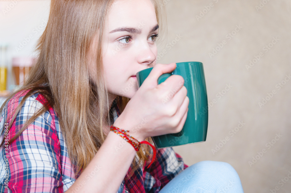 Attractive blonde girl is drinking tea from a large green mug in the kitchen. The concept of spending time during self-isolation of a virus pandemic Attractive blonde girl is drinking tea from a large green mug in the kitchen. The concept of spending time during self-isolation of a virus pandemic