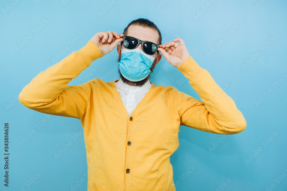 a man with glasses and a medical mask on a blue background. An emotional and positive guy on self-isolation. Coronavirus. Studio shooting. Pandemic COVID 19.