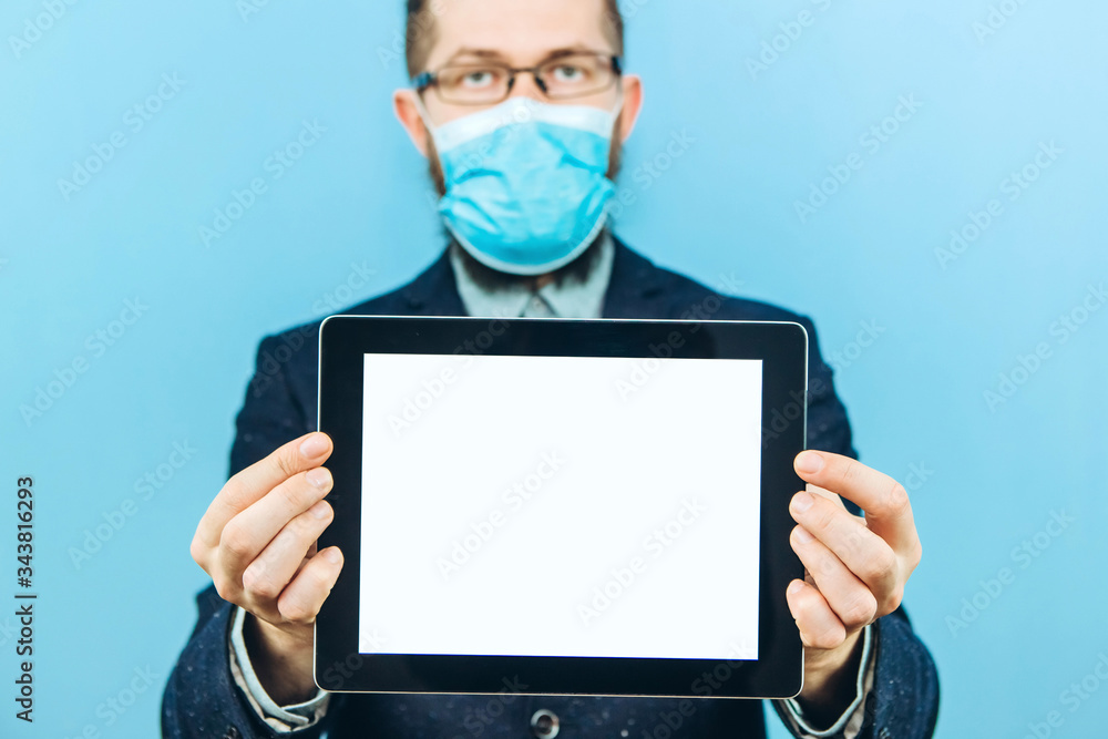 A man in a suit, glasses and a medical mask holds a piece of paper, on a blue background. Place for text. Businessman holds a tablet in his hands. The problem of business due to coronavirus. COVID-19 A man in a suit, glasses and a medical mask holds a piece of paper, on a blue background. Place for text. Businessman holds a tablet in his hands. The problem of business due to coronavirus. COVID-19
