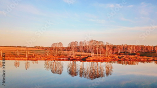 Frosty dry shore grass above calm river water reflecting blue morning sky, naked autumn trees