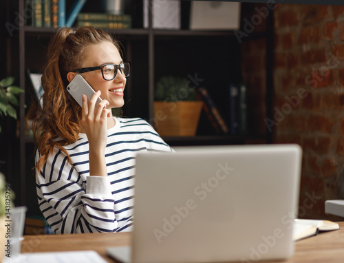 Young freelancer speaking on phone and works on a laptop in her home office.