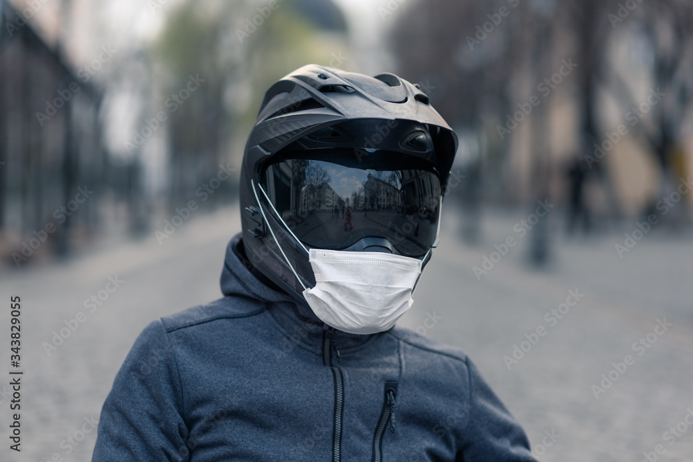 portrait of a man in a helmet. on a motorcycle helmet a white medical mask. photo on the street against the background of houses. coronavirus, disease, infection, quarantine, covid-19 portrait of a man in a helmet. on a motorcycle helmet a white medical mask. photo on the street against the background of houses. coronavirus, disease, infection, quarantine, covid-19