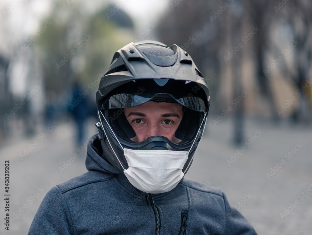 portrait of a man in a helmet. on a motorcycle helmet a white medical mask. photo on the street against the background of houses. coronavirus, disease, infection, quarantine, covid-19 portrait of a man in a helmet. on a motorcycle helmet a white medical mask. photo on the street against the background of houses. coronavirus, disease, infection, quarantine, covid-19