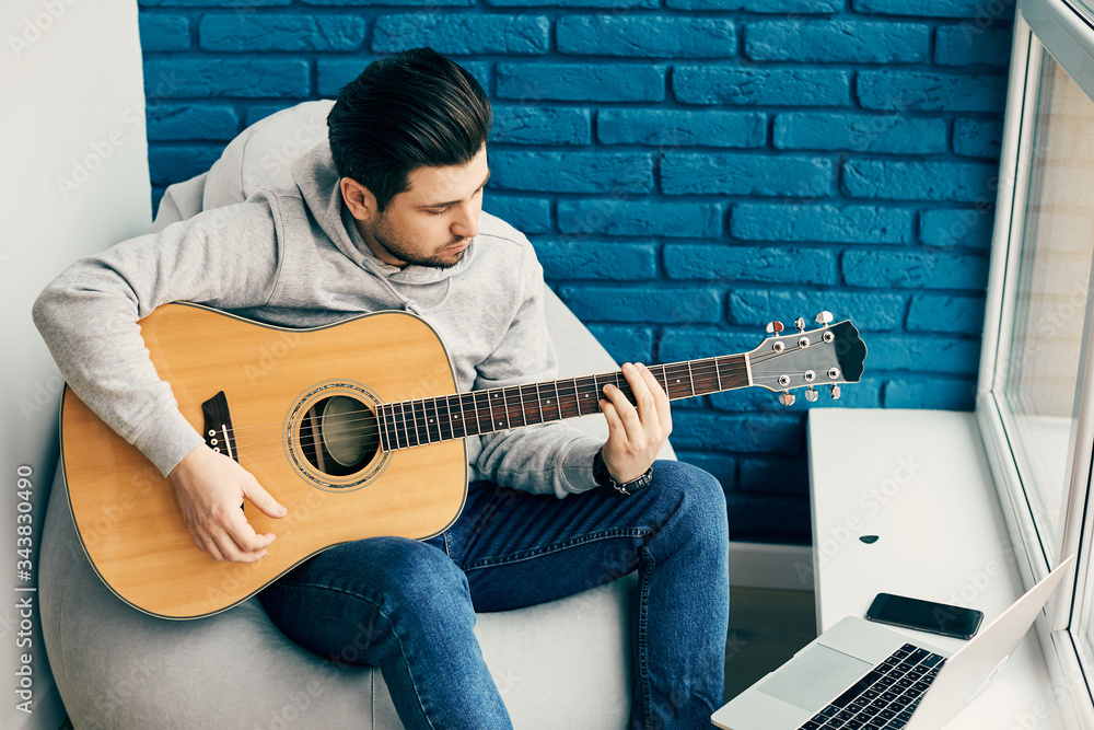 Obraz premium cheerful guitarist playing on instrument at home with laptop and phone on the side