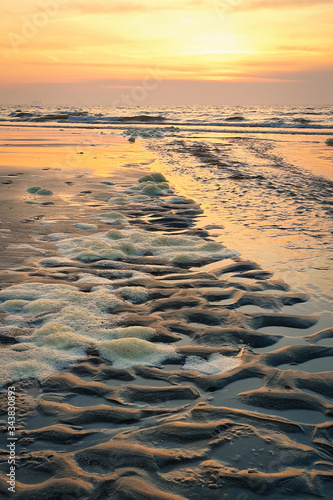 Beach with wave and ripple pattern in the sand is partly covered with foam. Sun is setting in the west.