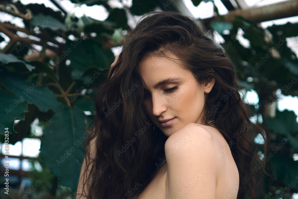 Fototapeta premium Portrait of a young brunette woman in orangery between green leaves; natural beauty, sunlight shining through the old greenhouse windows