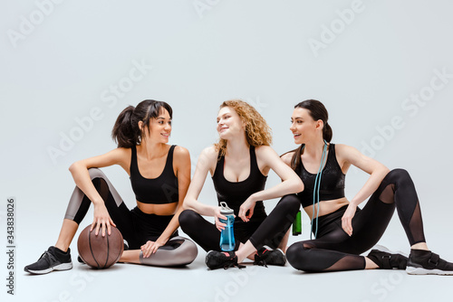 happy multicultural girls sitting with sport equipment on white