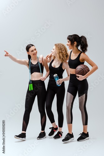 cheerful woman pointing with finger near multicultural girls walking with sport equipment on white