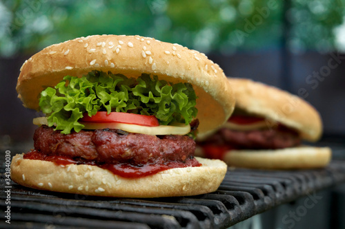 Tasteful white sesame bun burger standing on metal grill surface. Grilled beef on ketchup layer, cheese and tomato slices, leaf of crispy curly lettuce.Burgers on grill in garden party on summer day.