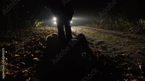 Handheld Shooting: The man beat the girl and drags her along the side of the road into the car at night on the background of rays of light from headlights photo