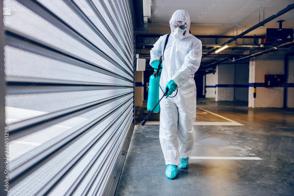 Full length of worker in sterile protective suit and mask sterilizing door of a garage from corona virus / covid-19. Full length of worker in sterile protective suit and mask sterilizing door of a garage from corona virus / covid-19.