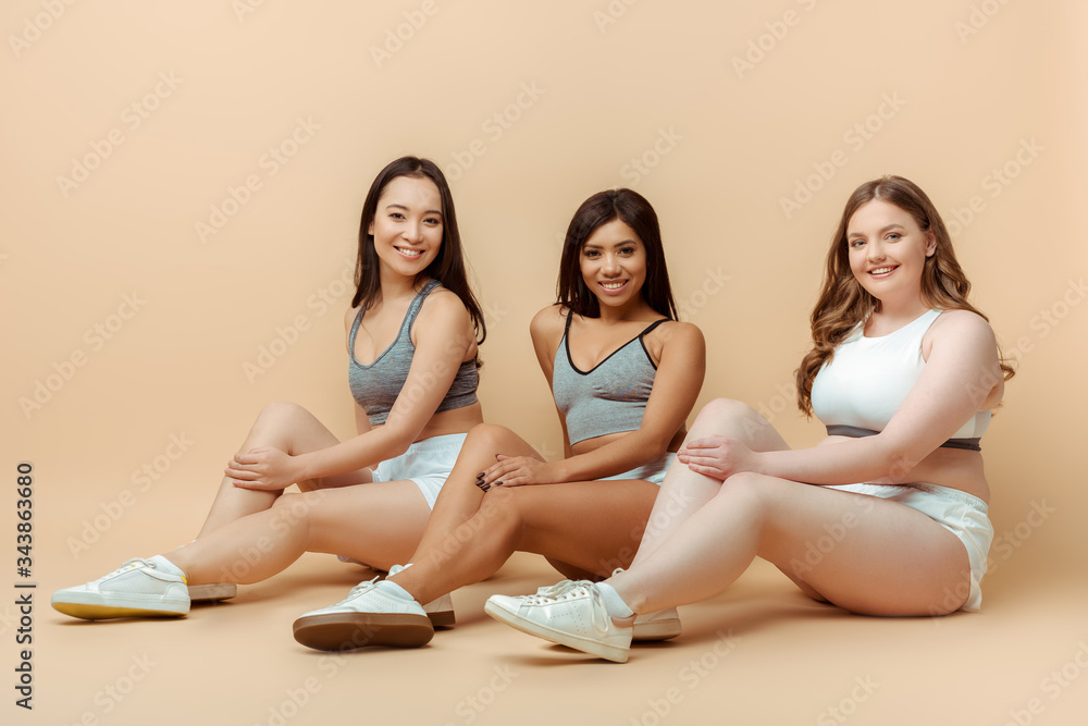 Multiethnic sportswomen sitting, smiling and looking at camera on beige