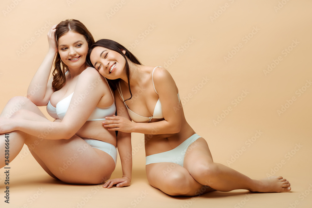 cheerful asian girl sitting with overweight woman in underwear on beige, body positive concept cheerful asian girl sitting with overweight woman in underwear on beige, body positive concept