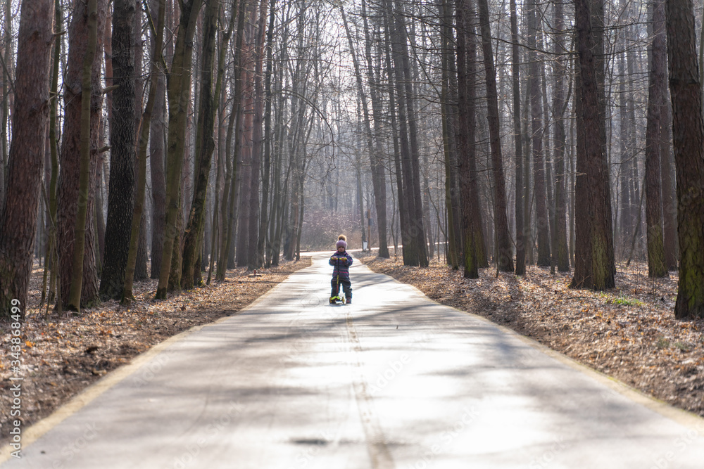 the child races on a scooter along an asphalt road through a spring park from the viewer. backlight