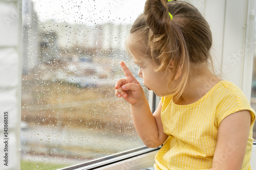 beautiful little girl looks out the window at the rain. stay home with coronavirus pandemic