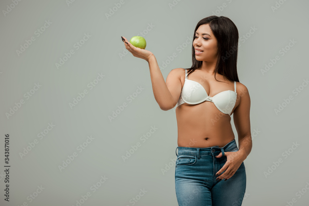 cheerful and overweight african american girl in jeans and bra holding apple isolated on grey cheerful and overweight african american girl in jeans and bra holding apple isolated on grey