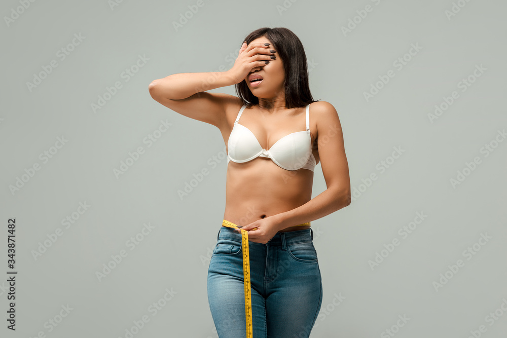 Naklejka premium overweight african american girl in jeans and bra measuring waist and covering eyes isolated on grey