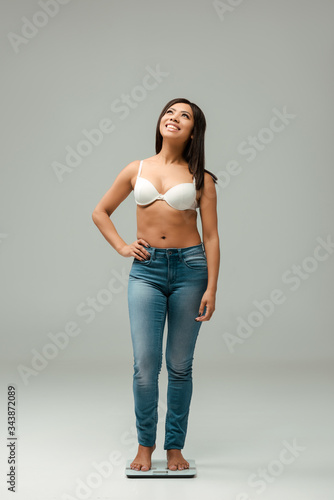 happy and overweight african american girl in jeans and bra standing with hand on hip on scales and looking up on grey