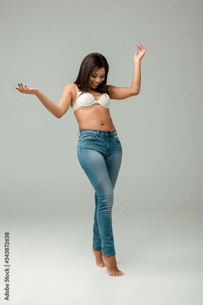 overweight and happy african american woman in jeans and bra standing on grey