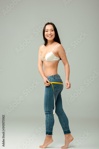 happy and overweight asian girl in bra measuring hips on grey
