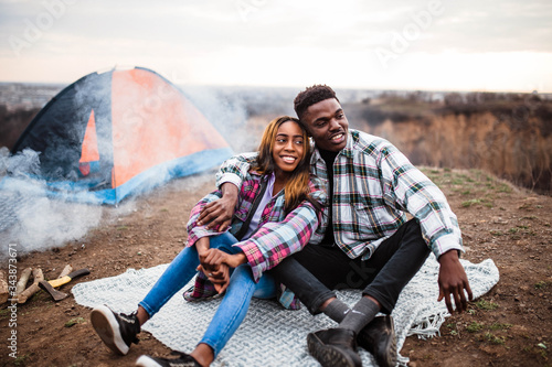 African American man and woman sitting on rug near campfire and tent, smiling, hugging each other. Man and woman in shirt.