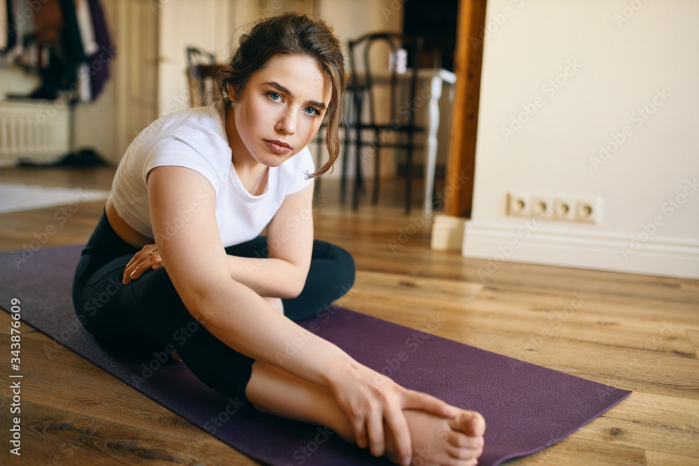 Serious frustrated young woman sitting on mat having painful look, suffering from injured leg because practicing yoga at home without proper alignment and supervision of professional instructor Serious frustrated young woman sitting on mat having painful look, suffering from injured leg because practicing yoga at home without proper alignment and supervision of professional instructor