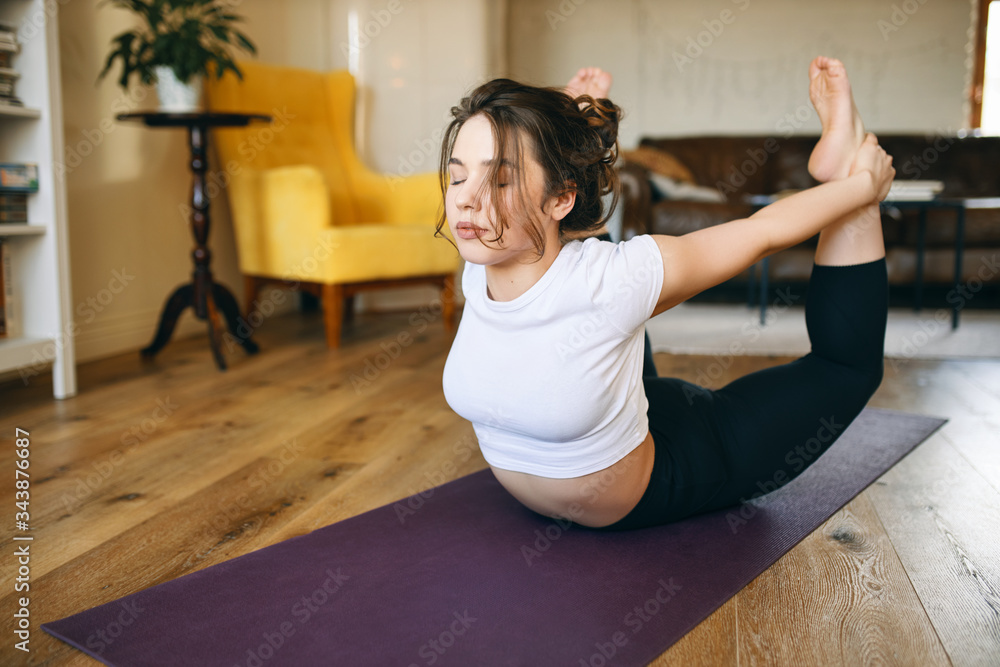 Flexible young female with curvy body exercising indoors, doing dhanurasana  or bow pose on yoga mat to strenghten back, open chest and shoulders,  improve digestion and stimulate reproductive organs Stock Photo