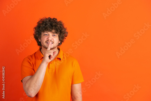 Whispering a secret. Caucasian young man's monochrome portrait on orange studio background. Beautiful male curly model in casual style. Concept of human emotions, facial expression, sales, ad.