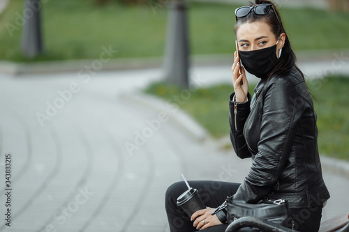 Portrait of brunette woman in a black protective mask sitting on a bench in the park, sunny spring day. Woman talking on mobile phone