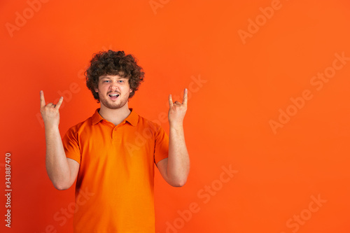 Rock horn gesture. Caucasian young man's monochrome portrait on orange studio background. Beautiful male curly model in casual style. Concept of human emotions, facial expression, sales, ad.