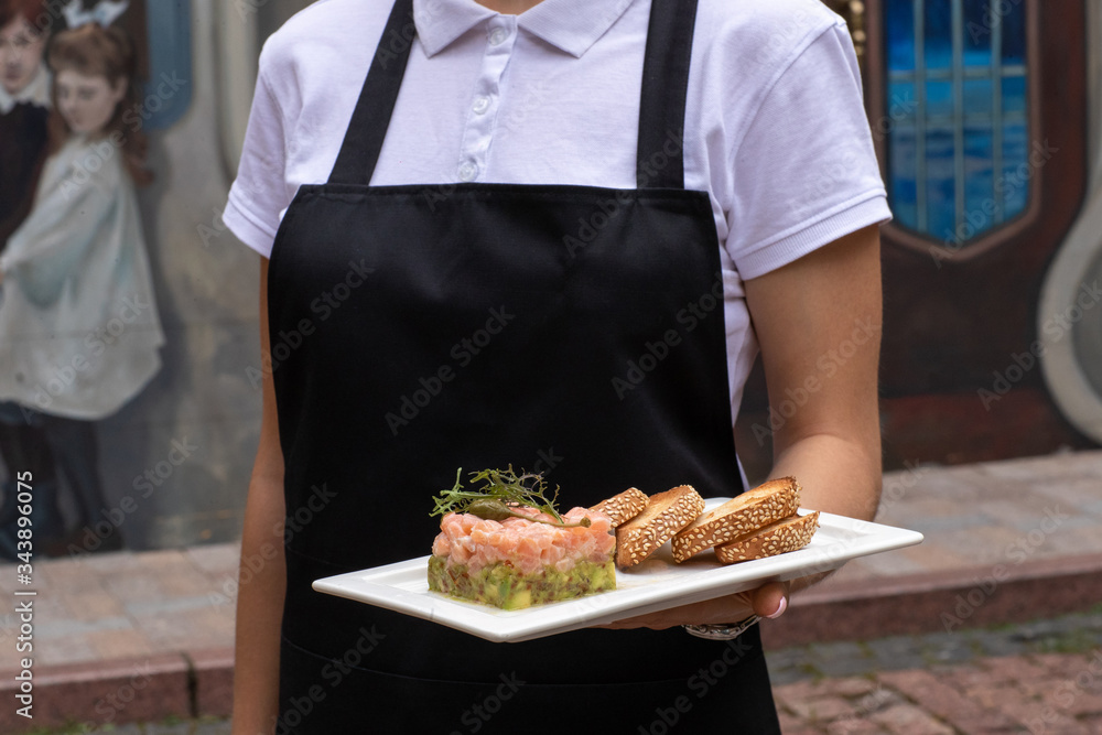 a girl in a black apron and a white bozka is holding in front of her a salmon avocado and rucola appetizer, .white bread cracker on a white plate. background street