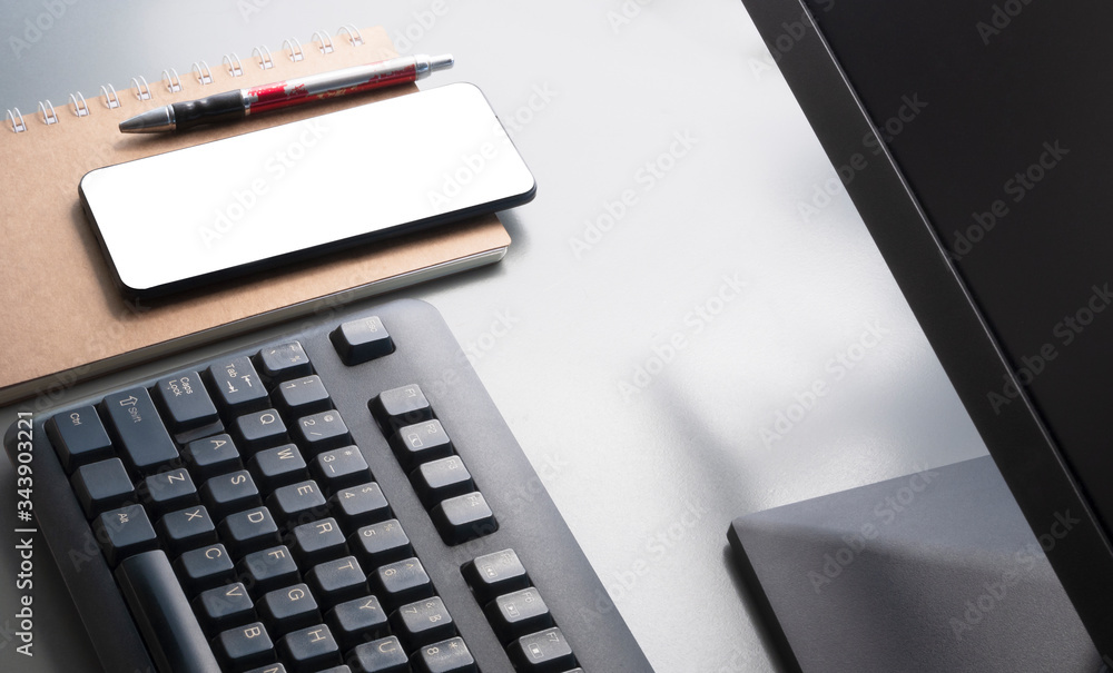 Morning soft light on surface of smartphone mock up, ballpoint pen, spiral notebook with part of computer keyboard and monitor on grey tabletop, high angle view and selective focus Morning soft light on surface of smartphone mock up, ballpoint pen, spiral notebook with part of computer keyboard and monitor on grey tabletop, high angle view and selective focus