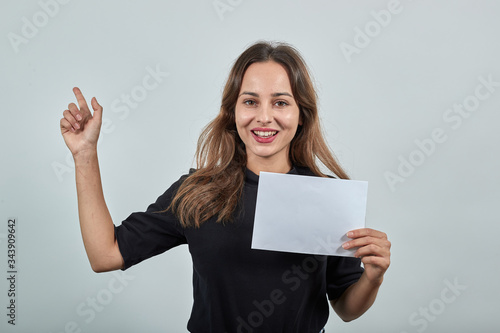 Cute young brunette woman in black t-shirt, blue jeans with belt on gray background, happy smiling girl holding a clean sheet of white paper in her hands and raised index finger up