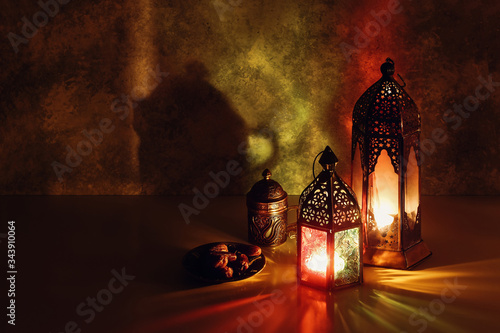 Festive greeting card, invitation for Muslim holiday Ramadan Kareem. Iftar dinner background. Decorative Arabic lanterns glowing at night, cup of tea, coffee and plate with dates on table. Shaby wall. photo