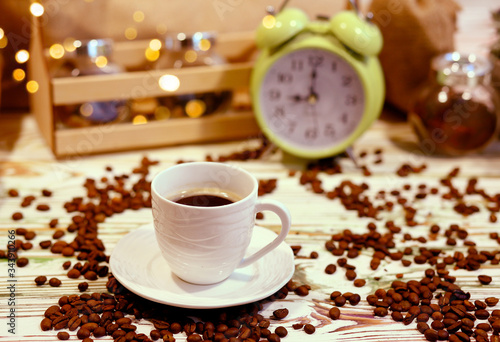 A cup of coffee on a platter with lots of sprinkled coffee beans. Alarm clock arrows at 9 am. Wooden textured table. Shy pastel background. Time to wake up in the morning. Natural food concept
