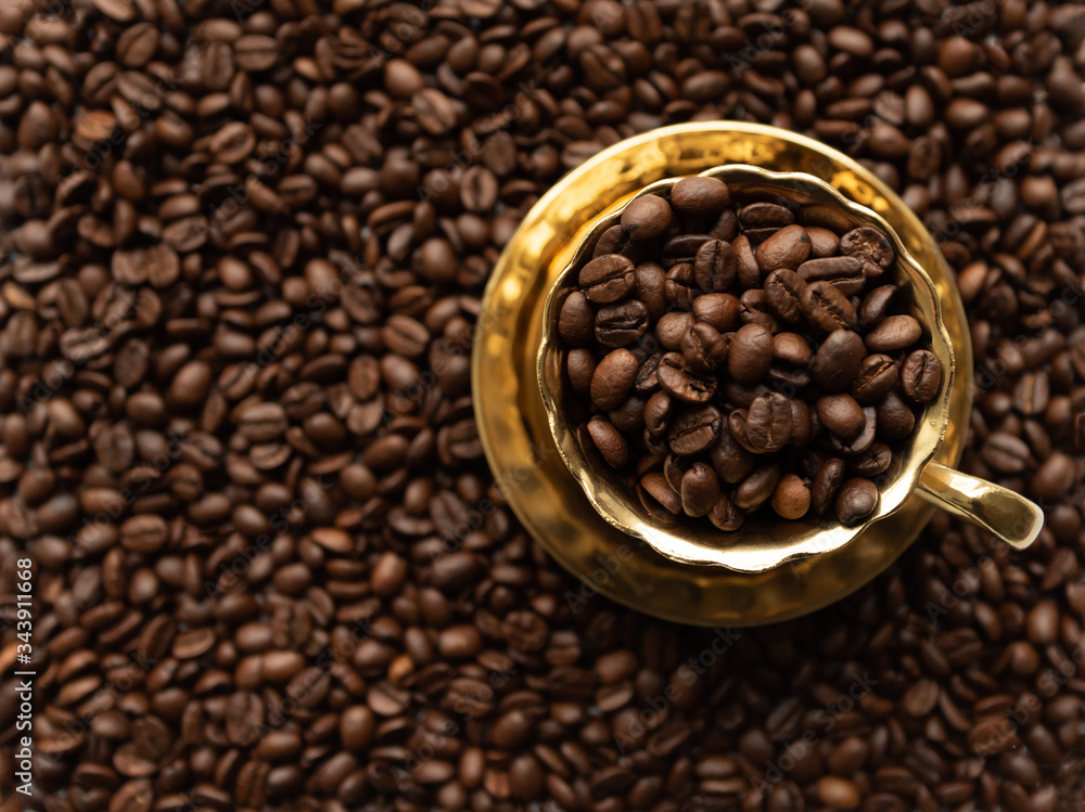 Golden cup on saucer filled with coffee beans on blurred background full of coffee beans