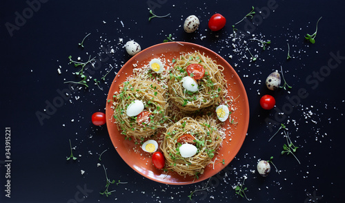 Buckwheat noodles with quail eggs, tomatoes and microgreen. Selective focus. Buckwheat pasta with cheese, vegetables and arugula in red plate on black background with ingredients. Horizontal, top view photo